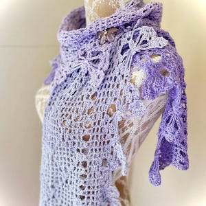 Puede incluir: Un chal de ganchillo hecho a mano, de color lavanda, con un diseño floral. El chal está drapeado sobre un maniquí, mostrando su intrincado calado y delicados bordes. Un broche decorativo está prendido al chal.
