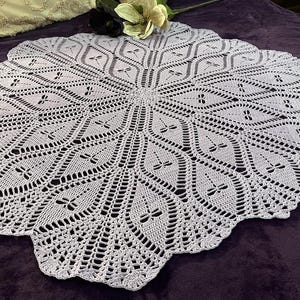 May include: A delicate, white crocheted tablecloth with a circular design featuring a repeating pattern of small, intricate flowers. The tablecloth has a scalloped edge and is laid out on a purple surface.