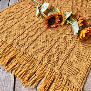 Puede incluir: Una manta de crochet de color amarillo mostaza con un borde de flecos. La manta está hecha con un patrón de puntada texturizado y está sobre una superficie de madera. Un ramo de girasoles está descansando sobre la manta.