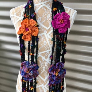 May include: A black crochet scarf with multiple strands and large, colorful crochet flowers. The flowers are in shades of orange, purple, and fuchsia. Small, colorful bobbles are scattered along the strands, adding texture and detail to the handmade accessory.