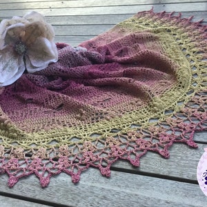A delicate shawl featuring dragonflies and a intricate lace edge, laid out on a deck, with a flower and a book.