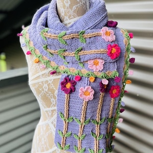 May include: A lilac crochet scarf adorned with floral and leaf details. The scarf showcases three-dimensional flowers in pink and purple hues, complemented by green leaves and beige stems. The edges are finished with small, colourful bobbles.