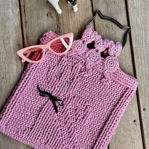 May include: A pink crocheted tote bag with a cat-shaped handle and black ribbon bow. The bag has a crocheted cat design on the front and two rhinestones.