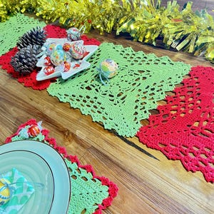 May include: A festive Christmas table setting featuring a green and red crocheted table runner. The runner is adorned with a Christmas tree-shaped dish filled with decorative candies, pine cones, and a gold tinsel garland.