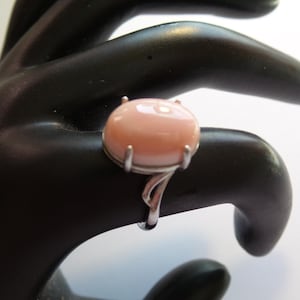 May include: A silver ring featuring a large, oval, light pink gemstone. The gemstone is set in a silver band with a decorative, swirling design on the sides. The ring is displayed on a black hand.