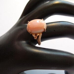 May include: A rose gold ring featuring a large, oval-shaped, peach-colored gemstone. The ring has a delicate, ornate design with small, sparkling accent stones. The ring is displayed on a black surface.