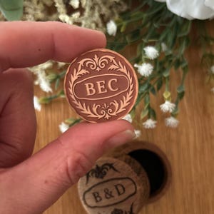 May include: A round, brown medallion with the letters "BEC" in a decorative frame is held in a person's hand. Below, a small, round box with the letters "B&D" is visible. The medallion and box appear to be made of wood.