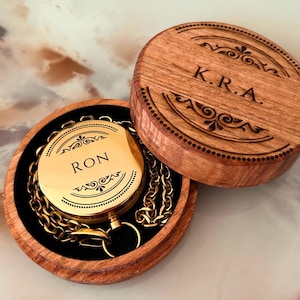 May include: A gold pocket watch, engraved with the name "RON", inside a wooden box. The box lid is engraved with the initials "K.R.A." and decorative patterns. A gold chain is attached to the watch. The box is made of wood.