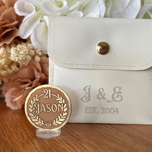 May include: A cream-colored pouch with gold hardware and the engraved initials "J & G" and "EST. 2004". A bronze-colored coin with the number "21" and the name "JASON" is in front of the pouch. The background features flowers.