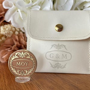 May include: A cream-colored pouch with a gold button closure and the initials "G & M" is next to a round, brown and gold decorative item with the name "MOY". The items are set against a backdrop of flowers.