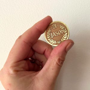 May include: A round, bronze-colored coin with the name "JASON" and the number "21" engraved on it. The coin is held between fingers against a white background. The coin has a gold-colored border and laurel wreath design.