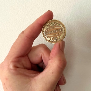 May include: A round, gold-colored coin with the word "GEORDIE" in a decorative oval. The coin has an ornate, leaf-like border. The coin is held between fingers against a white background.