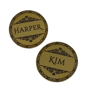 May include: Two round, gold-colored metal name tags with decorative borders and black engraved lettering. The name tags read "HARPER" and "KIM".