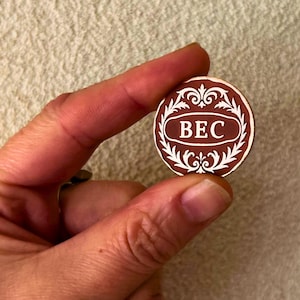 May include: A round, brown and white decorative token with the letters "BEC" in the center. The token has a decorative border and is held between two fingers.