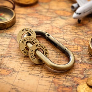 May include: A brass-toned metal carabiner keychain featuring three circular brass rings attached to the loop. Each ring is stamped with text including NEPAL, PHILIPP, MONTENE, 2005, and 2019. The carabiner has a spring-loaded gate mechanism.