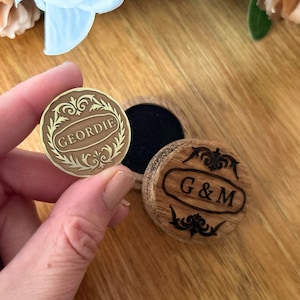 May include: A small, round, wooden box with a hinged lid, and a separate coin. The coin is held in a hand and has the name "GEORDIE" engraved on it. The box lid has the initials "G & M".