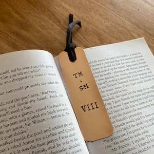 May include: A close-up of a brown leather bookmark with the initials "TM + SM VIII" engraved on it. The bookmark is lying on top of an open book.