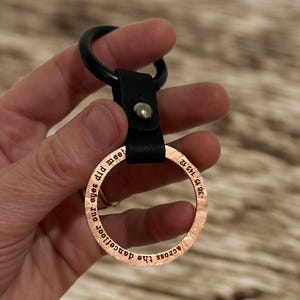 Könnte beinhalten: Ein kupferfarbener Metall-Schlüsselanhänger mit den Worten "across the dancefloor our eyes did meet" am Rand eingraviert. Der Schlüsselanhänger hat einen schwarzen Lederriemen und einen schwarzen Metallring.