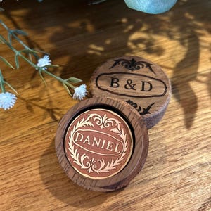May include: Two wooden coasters with engraved designs. One coaster features the initials "B & D", while the other displays the name "DANIEL" in an ornate frame. The coasters are set on a wooden surface.