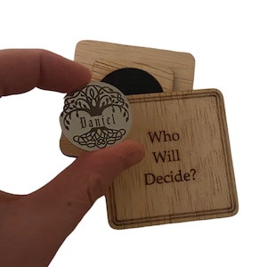 May include: A wooden box with the text "Who Will Decide?" on the front. The box is open and a silver coin with a tree design and the name "Daniel" is visible.