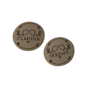 May include: Two round, taupe-colored coasters with a decorative design. Each coaster features a floral and heart border, with the names "CLAUDIA" and "JEREMY" in a script font. The coasters are set against a white background.