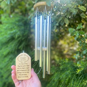May include: A wooden wind chime with silver tubes and a wooden plaque that reads "DAD 1947-2020 In loving memory May these gentle chimes echo the whispers of your spirit, bringing peace and serenity to all who listen. Rest peacefully forever in our hearts."