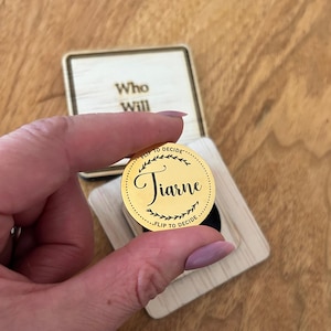 May include: A gold coin with the name "Tiare" inscribed in a circle with a floral design. The coin is on a wooden box with the text "Who Will Flip to Decide".
