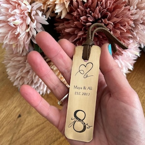 May include: A wooden bookmark with a brown leather strap. The bookmark is engraved with a heart, the text "Maya & Ali EST. 2017", and the number "8".