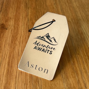 May include: A rectangular, light-colored tag with a black string, featuring the text "Adventure Awaits" above the name "Aston" in a sans-serif font. The tag has a mountain graphic and is set against a wooden background.