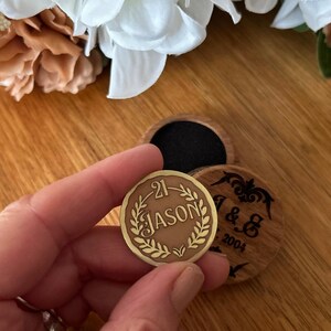 May include: A round, bronze-colored coin with the name "JASON" and the number "21" engraved within a laurel wreath. The coin is held near a wooden box with a black interior and a wooden lid with the initials "J & G" and the year "2004".
