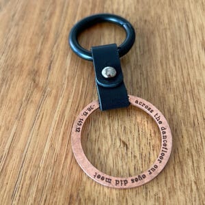 Könnte beinhalten: Ein kupferfarbener Schlüsselanhänger mit graviertem Text, verbunden mit einem schwarzen Lederriemen und einem schwarzen Metallring. Der Text lautet "...our eyes did meet across the dancefloor..." und enthält Koordinaten. Der Schlüsselanhänger liegt auf einer Holzoberfläche.