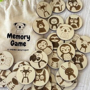 Puede incluir: Un juego de memoria de madera con ilustraciones de varios animales. Las fichas son de color marrón claro con detalles en negro y marrón. Las fichas están dispuestas en círculo sobre un fondo blanco. El texto "Memory Game" está impreso en una pequeña bolsa de lona en el fondo.