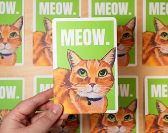 May include: A rectangular card with a green border and the word "MEOW" in white. The card features an orange cat with green eyes and a black collar. Other cards with the same design are in the background.