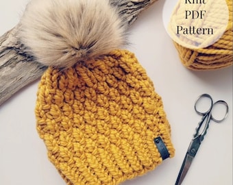 May include: A mustard yellow knit beanie with a fluffy brown pom pom. The beanie has a black leather tag with the word "UTW" on it. The beanie is on a white background with a ball of yarn and scissors.