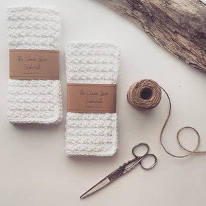 May include: Two white linen dishcloths with brown labels that read "The Classic Linen Dishcloth". The dishcloths are on a white surface with a piece of driftwood, a ball of twine, and a pair of scissors.