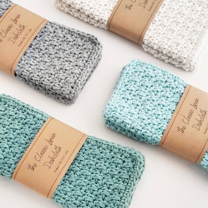 May include: Four crocheted dishcloths in various colours, including grey, white, and two shades of blue. Each dishcloth has a brown paper tag with the text "The Classic Linen Dishcloth" and "Handmade with 100% Cotton".