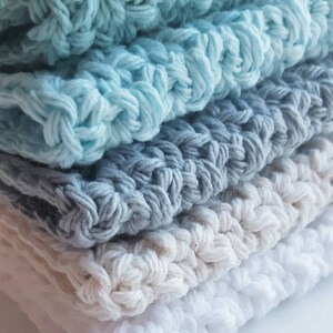 May include: Stack of four crocheted dishcloths in shades of blue, grey, and white. The cloths are made with a simple stitch pattern and have a soft, textured feel.
