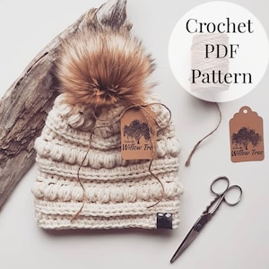 May include: A crocheted beanie hat with a fluffy brown pom pom. The hat is cream colored and has a textured stitch pattern. The hat is tied with a brown string and has a brown tag that says "Under the Willow Tree". The image also includes a ball of yarn, a pair of scissors, and a second brown tag with the same text.