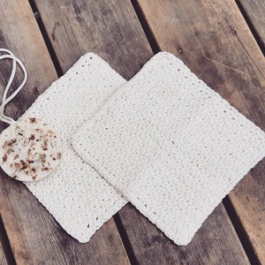 May include: Two white crocheted dishcloths or scrubbers, made with a textured stitch, are laid out on a wooden surface.