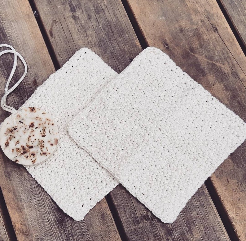May include: Two white crocheted dishcloths or scrubbers, made with a textured stitch, are laid out on a wooden surface.