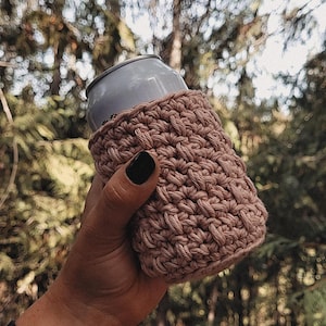 May include: A hand-crocheted, light brown can cozy. The cozy is wrapped around a silver can, held against a blurred background of green trees. The cozy is made of a thick, textured yarn.