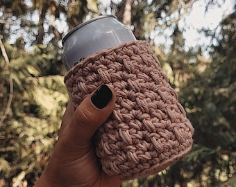 May include: A hand-crocheted, light brown can cosy. The cosy is wrapped around a silver can, held against a blurred background of green trees. The cosy is made of a thick, textured yarn.
