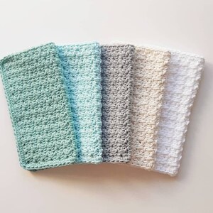 May include: Five crocheted dishcloths in different colours: light green, light blue, grey, beige, and white. The dishcloths are stacked on top of each other.