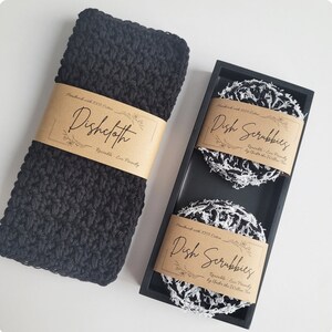 May include: A set of two black and white dish scrubbies and a black crocheted dishcloth. The scrubbies and dishcloth are handmade with 100% cotton and are reusable and eco-friendly. The scrubbies are packaged in a black wooden box with a label that reads "Dish Scrubbies Handmade with 100% Cotton Reusable - Eco Friendly by Under the Willow Tree". The dishcloth has a label that reads "Dishcloth Handmade with 100% Cotton Reusable - Eco Friendly".