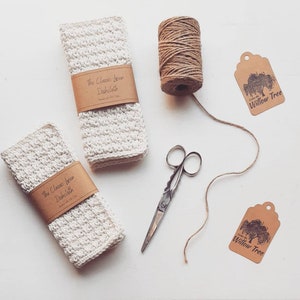 May include: Two white linen dishcloths with the text "The Classic Linen Dishcloth" printed on brown paper labels. The dishcloths are on a white surface with a spool of twine, scissors, and two brown paper tags with the text "Willow Tree" printed on them.