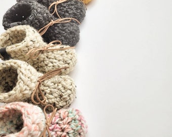 May include: A row of crocheted baby booties in various colours, including grey, white, beige, yellow, and pink. The booties have brown leather ties.