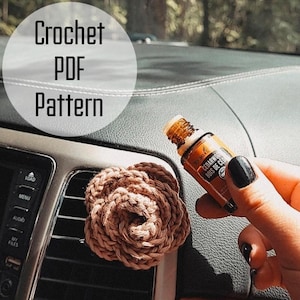Puede incluir: Un ambientador de flor de ganchillo marrón sujeto a la rejilla de ventilación de un coche, con un pequeño frasco de vidrio ámbar de aceite esencial de cedro cerca. El texto Crochet PDF Pattern aparece en un círculo blanco a la izquierda.