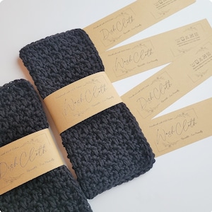 May include: Four brown paper tags with the text "Wash Cloth" and "Dish Cloth" in black cursive writing. The tags are attached to three black crocheted dishcloths.