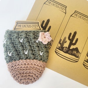 Op de afbeelding: Een groene en bruine gehaakte cactus-blikjeskoeler met een roze bloem aan de zijkant. De koeler heeft de tekst "The Cactus Cozy" aan de voorkant.