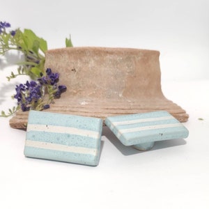 Può includere: Due pomelli quadrati in ceramica azzurra con strisce bianche. I pomelli sono esposti su una superficie bianca con un pezzo di ceramica marrone usurato sullo sfondo. Sono visibili anche fiori viola e foglie verdi.
